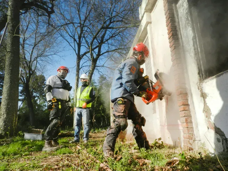 Three people in safety gear near a building; one cuts the wall with a power saw.