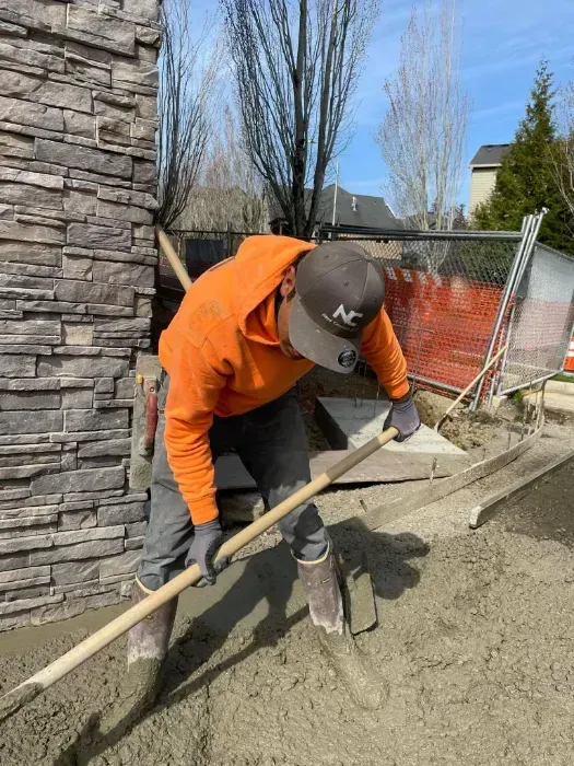 Construction worker in orange hoodie leveling wet concrete with a rake outside near a stone wall and fence.