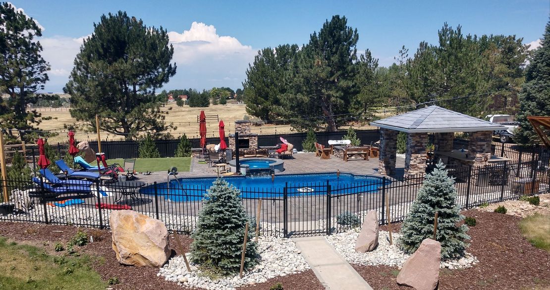A backyard with a pool covered by a blue tarp, surrounded by a black fence and landscaping. Trees and a stone patio are also visible.
