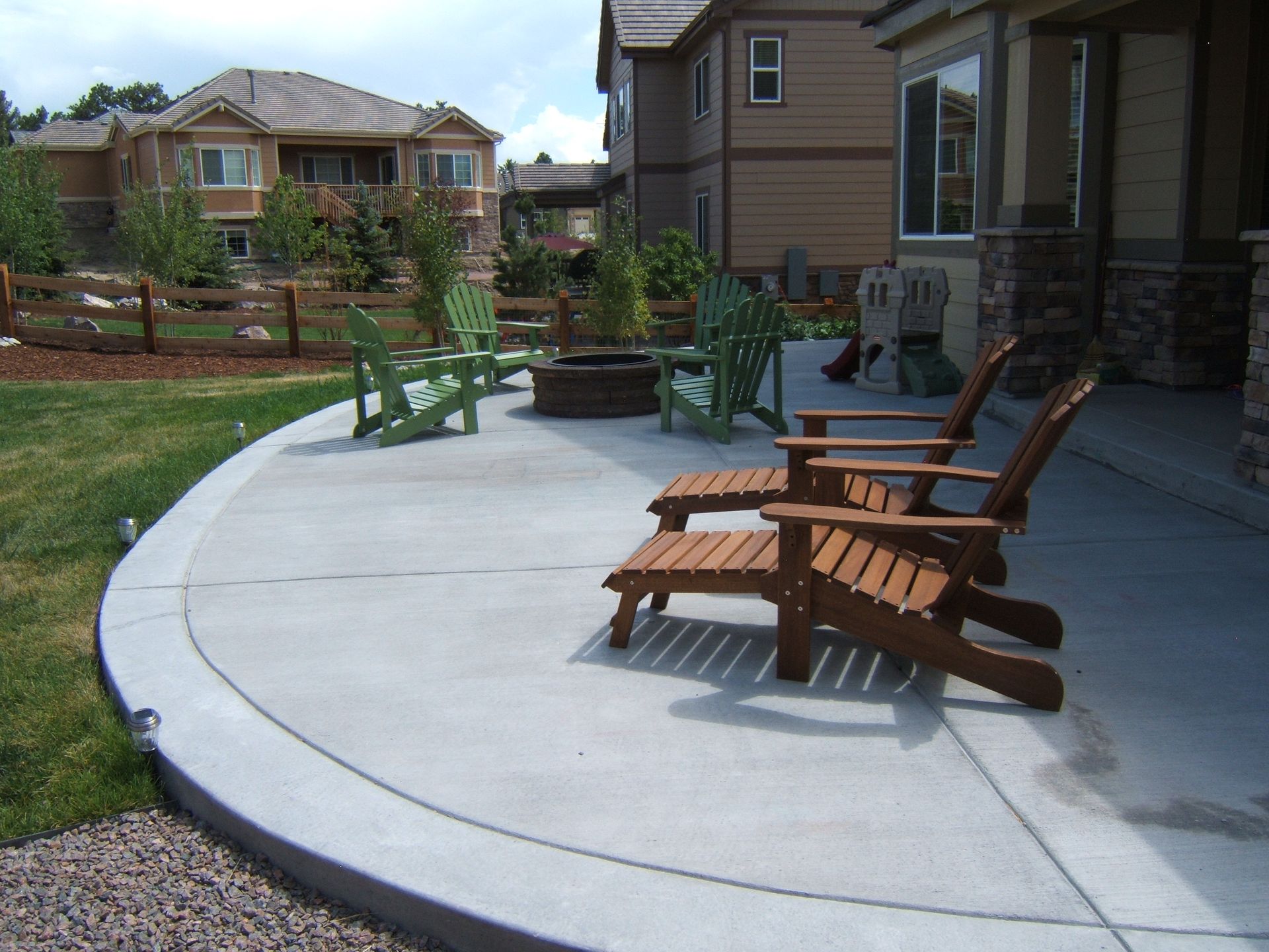 Patio with wooden chairs, including green Adirondack chairs, near a house.