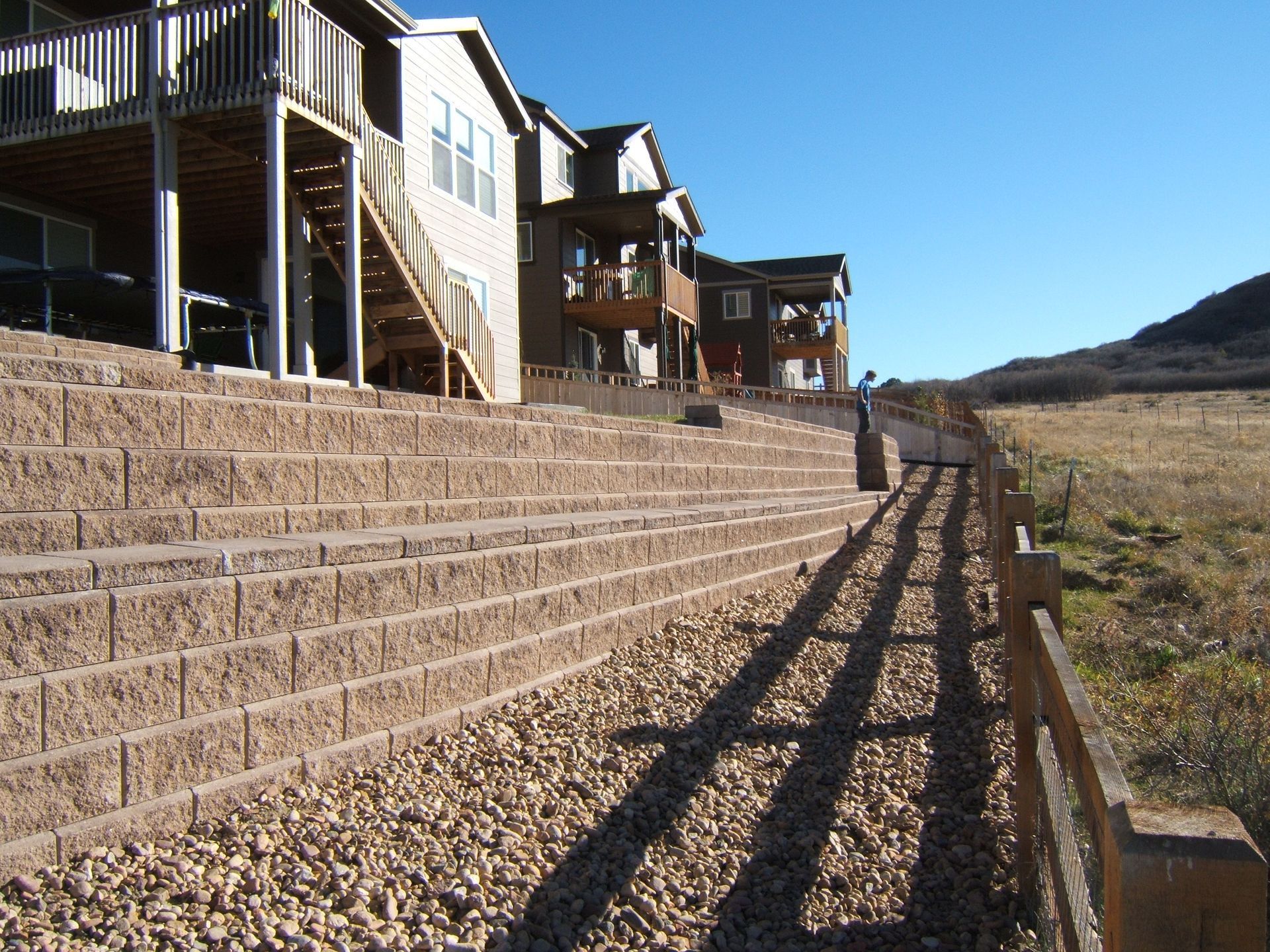 Brown retaining walls supporting homes on a hillside next to a wooden fence and grassy field.