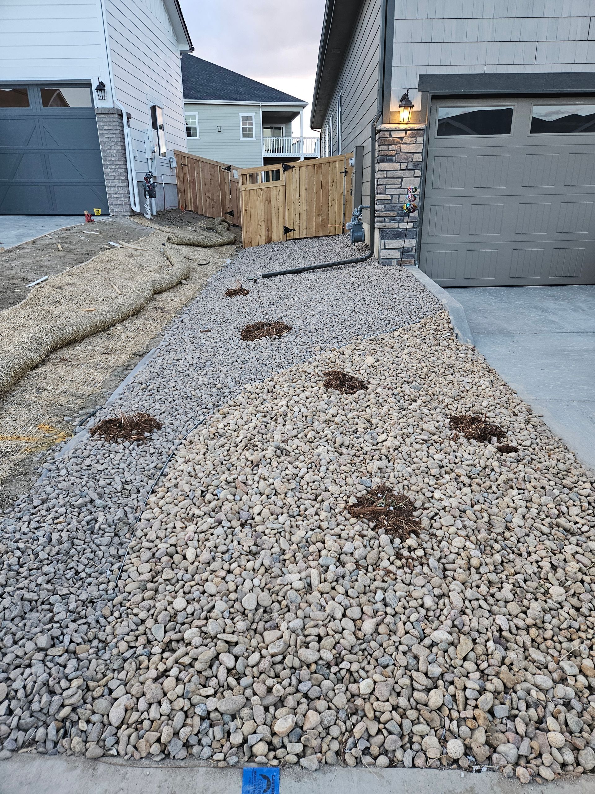 A gravel driveway with several planting areas leads to a garage. A wooden fence and building are visible in the background.
