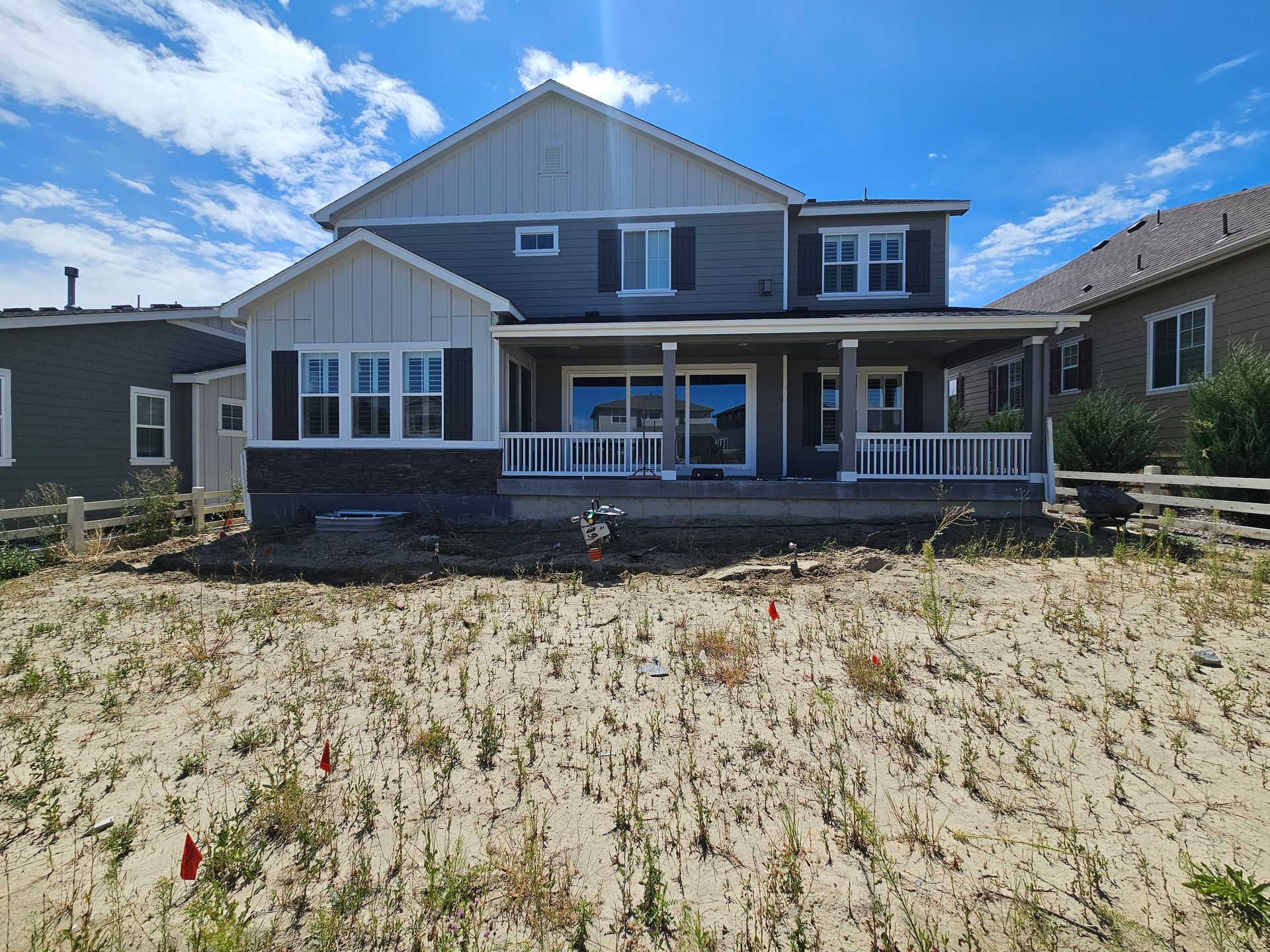 Back view of a gray two-story house with a covered porch, set against a blue sky with clouds. The yard is dry with sparse vegetation.