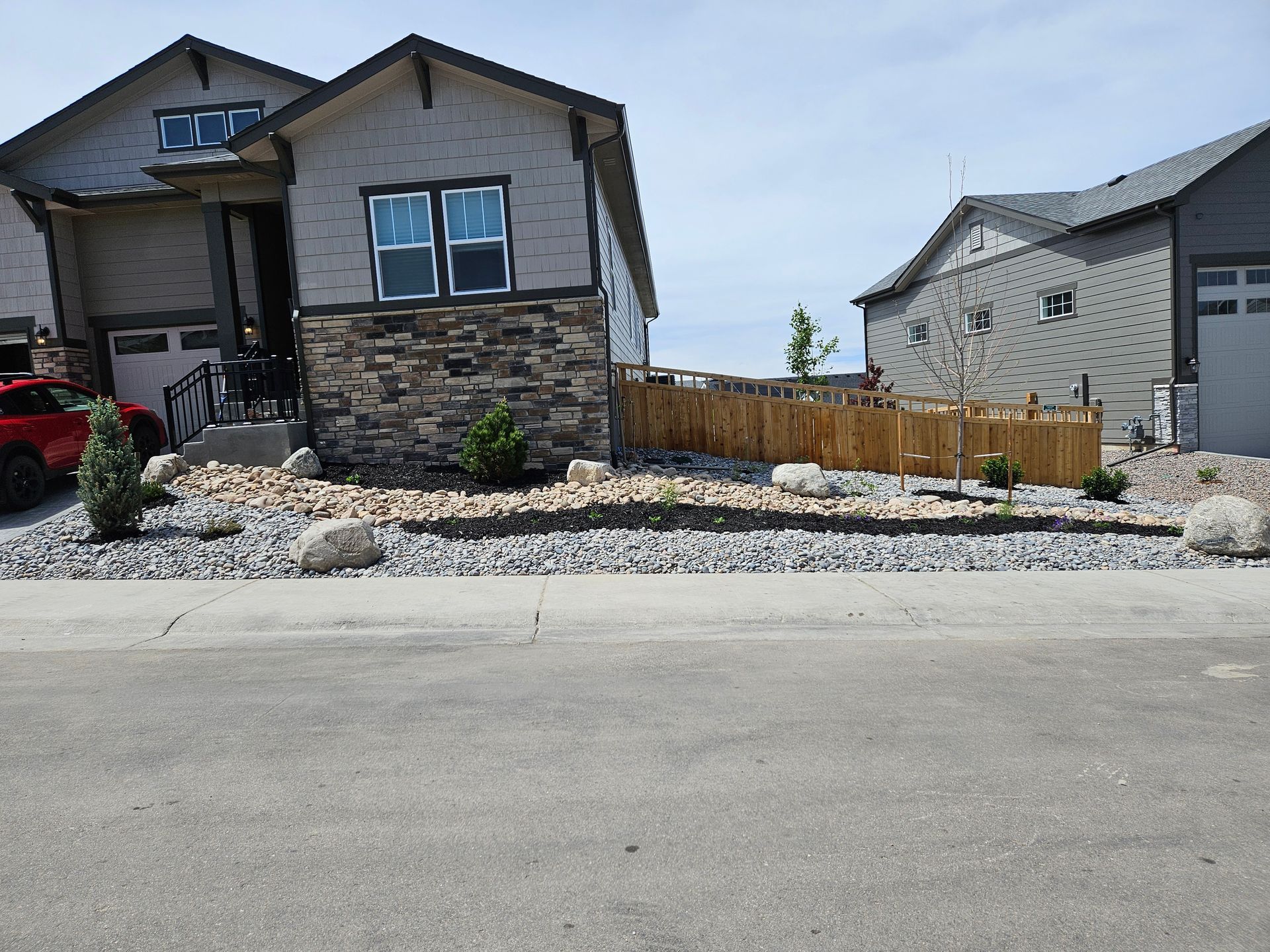 A modern house with stone and gray siding, a rock garden, and a wooden fence on a sunny day.