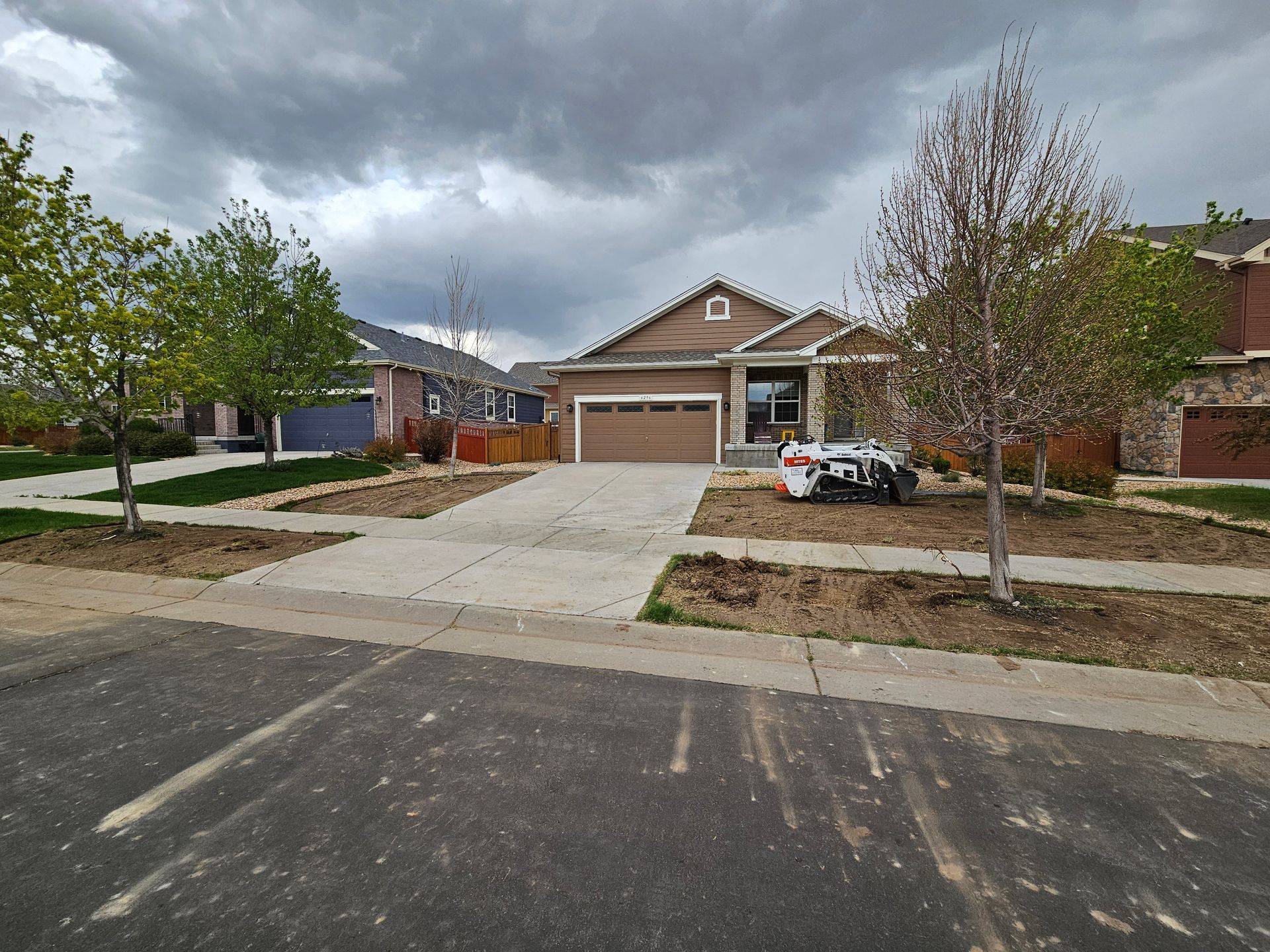 A light brown house with a two-car garage under a cloudy sky. Front yard features small trees and empty planting beds.