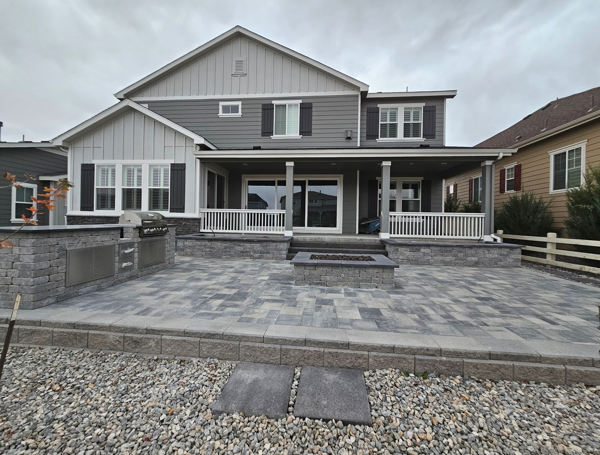 Backyard patio with gray pavers and a fire pit, leading to a house with a covered porch. Overcast sky.