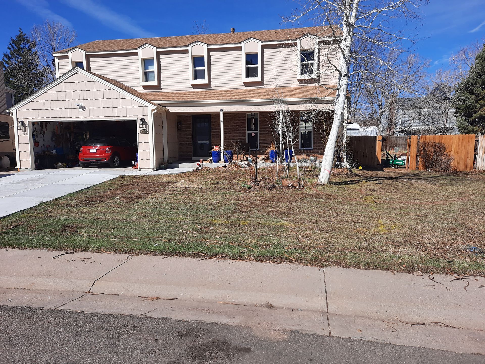 Two-story beige house with a garage, porch, and yard. A red car is in the garage.  A blue sky is overhead.