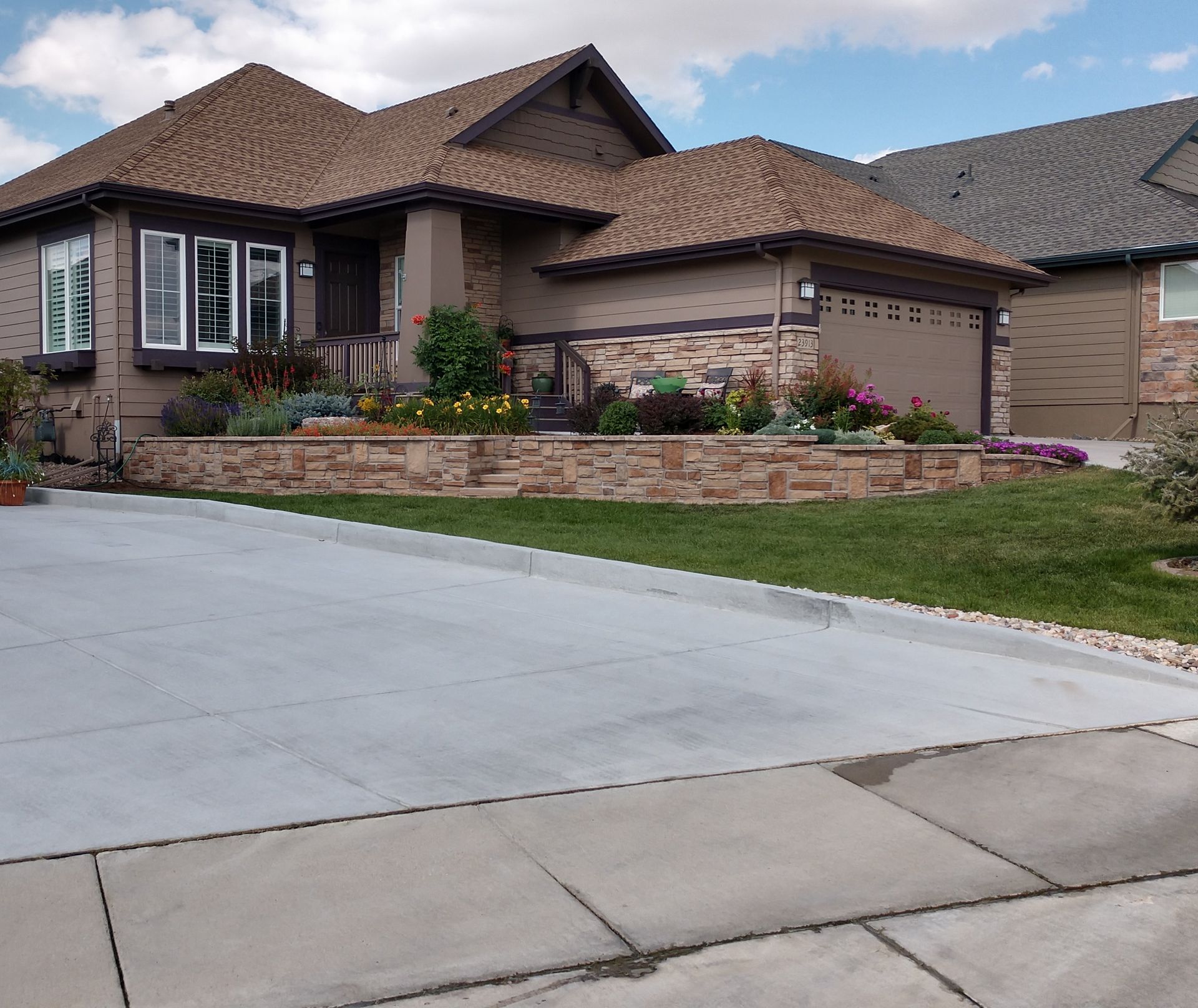 A tan house with a stone-faced retaining wall and colorful flowerbeds. A gray driveway and sidewalk are in the foreground.