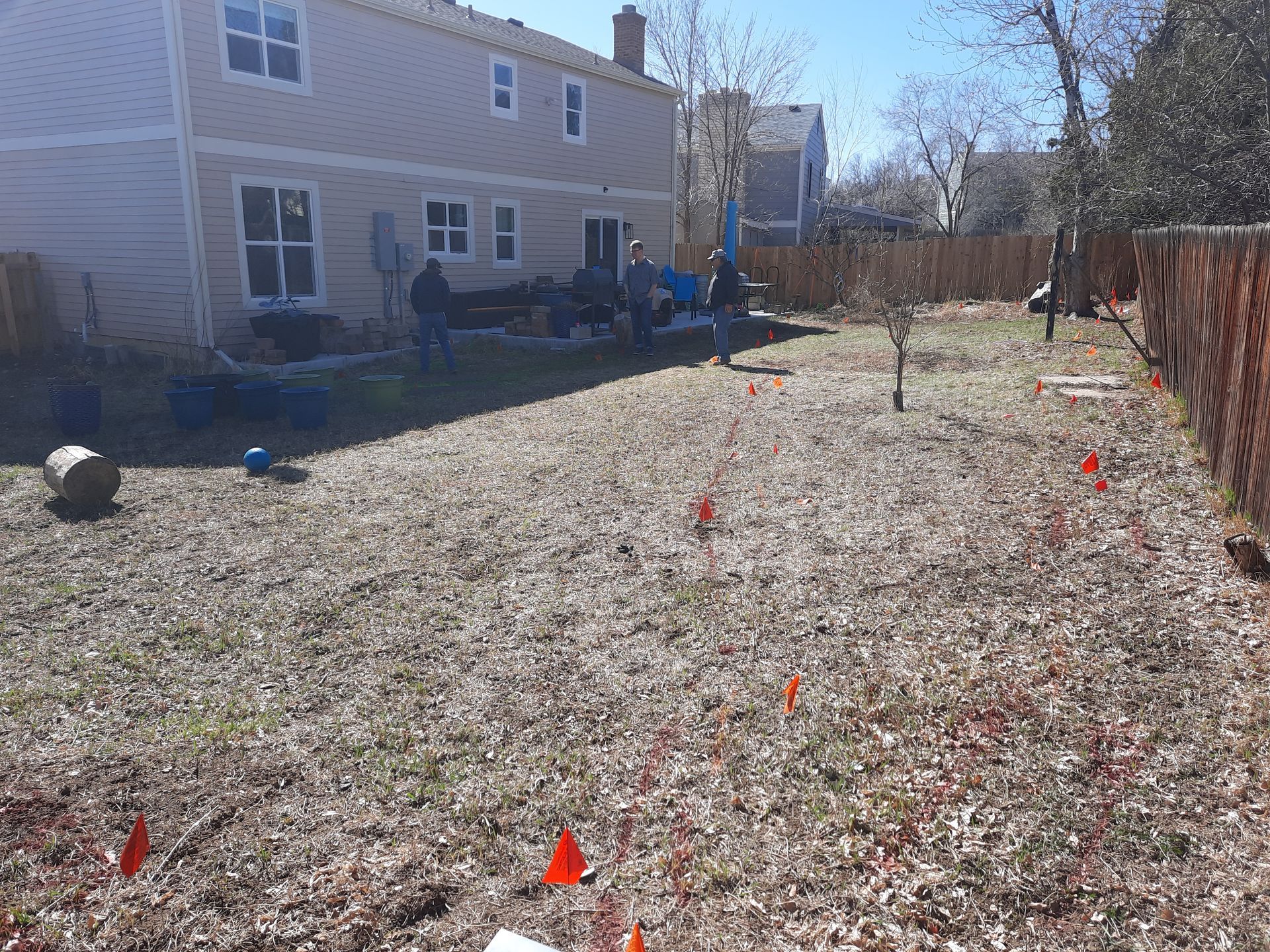 Backyard with sparse grass marked with orange flags, near a wooden fence and a two-story house. People are in the background.