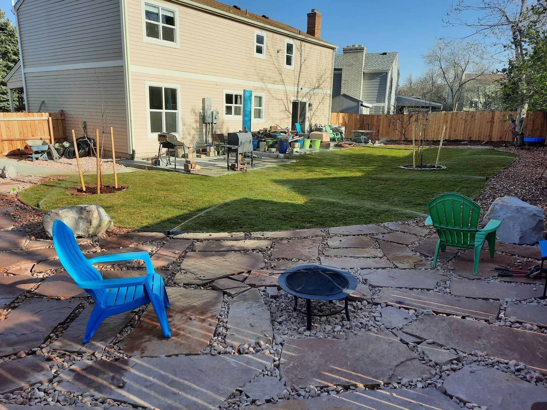 Backyard with a flagstone patio, fire pit, blue and green chairs, and a grassy lawn leading to a two-story house with a wooden fence.