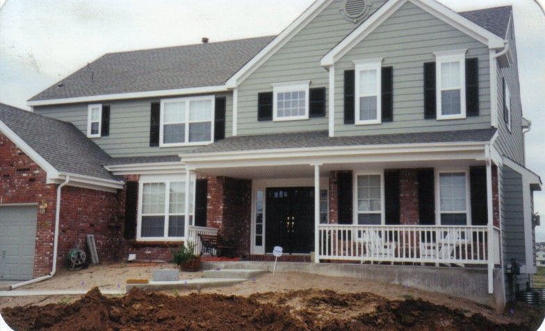 Two-story house with green siding, brick accents, and black shutters. A covered porch has white railings and a white trim.