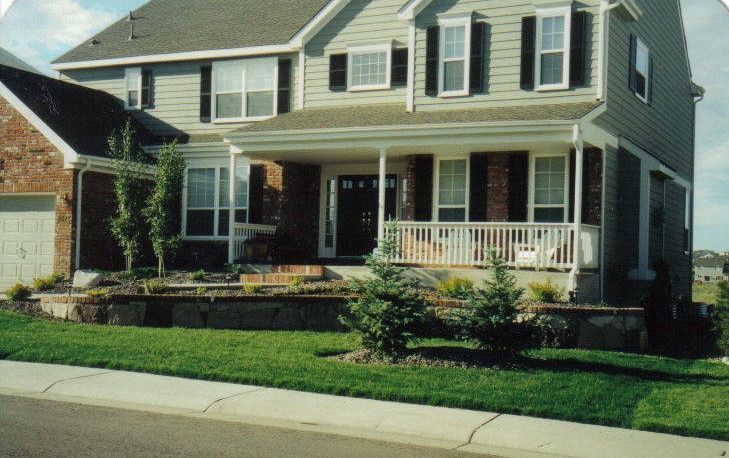 Two-story house with a porch. Tan siding, red brick accents. Green lawn, trees, and sidewalk in front.