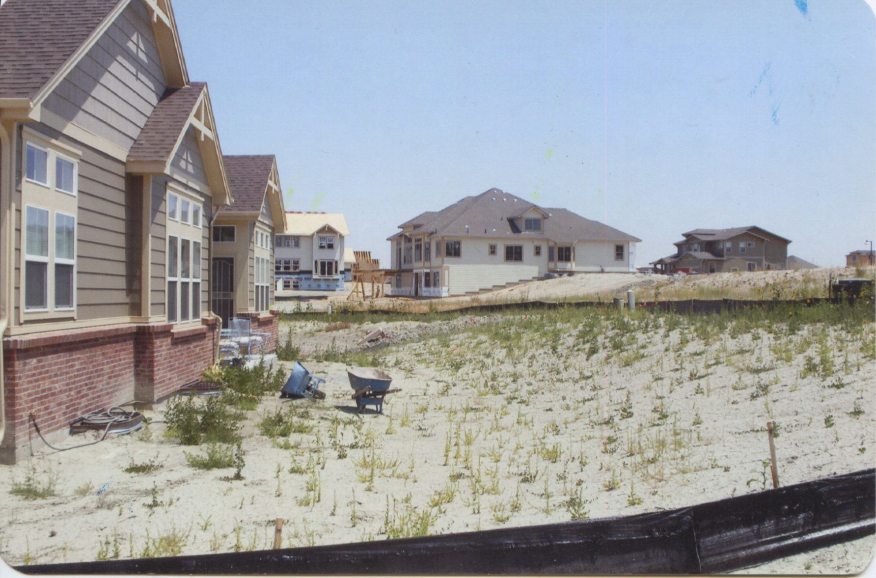 Houses under construction on sandy land with some weeds; two people work near a wheelbarrow on the sandy ground.