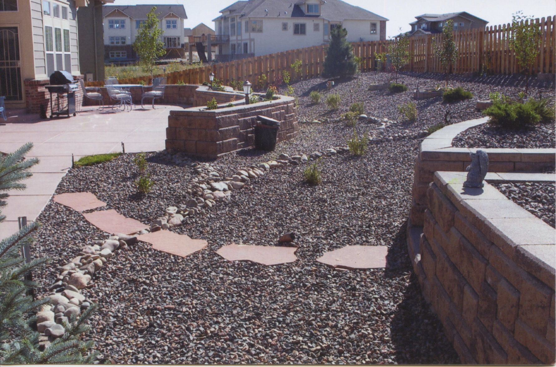 Backyard patio with brick retaining walls, stone pathway, and gravel landscaping; a barbecue and seating are on the patio.