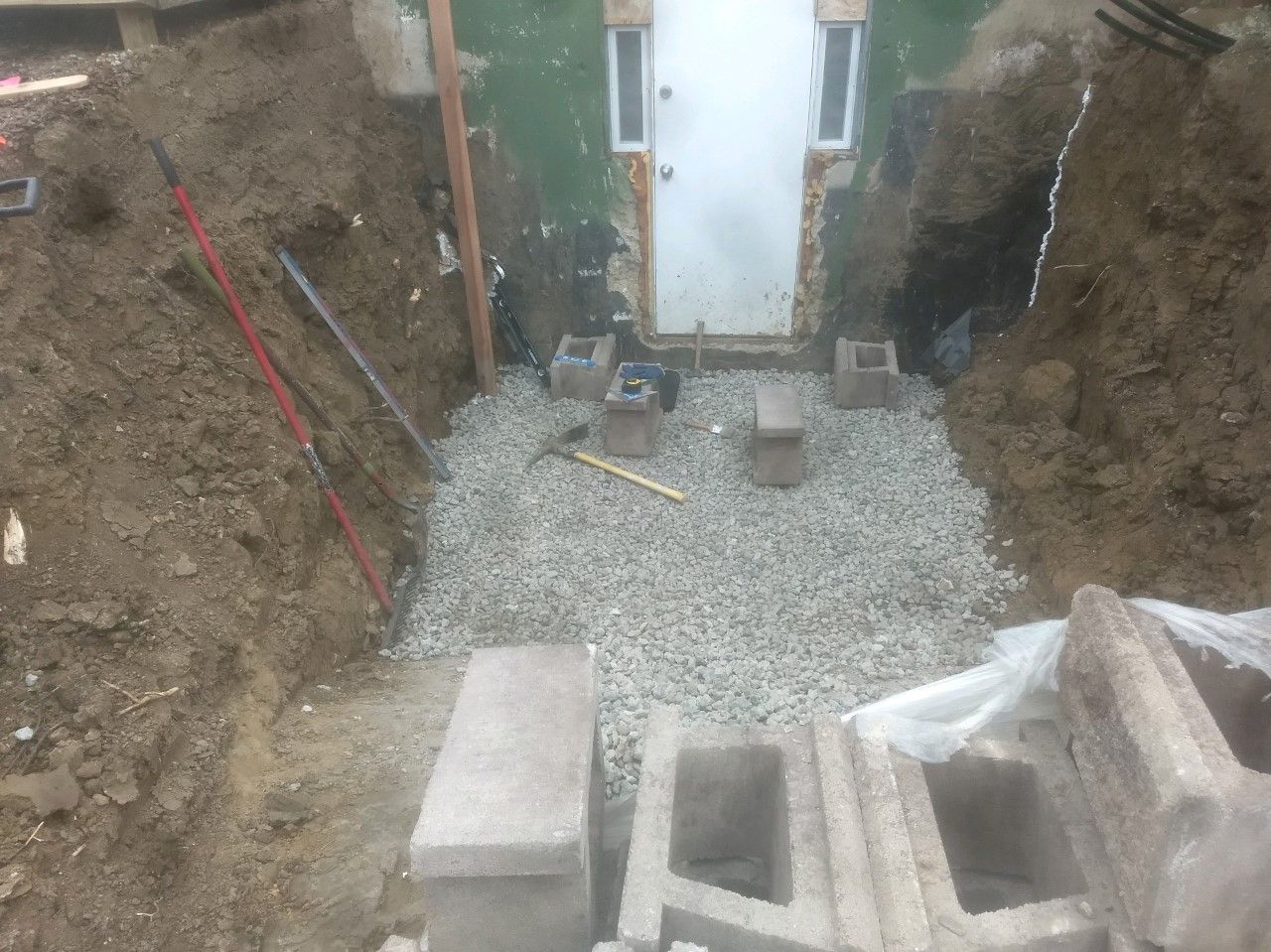 An excavated doorway surrounded by dirt, leading to a white door. The area is covered in gravel with concrete blocks placed around it.