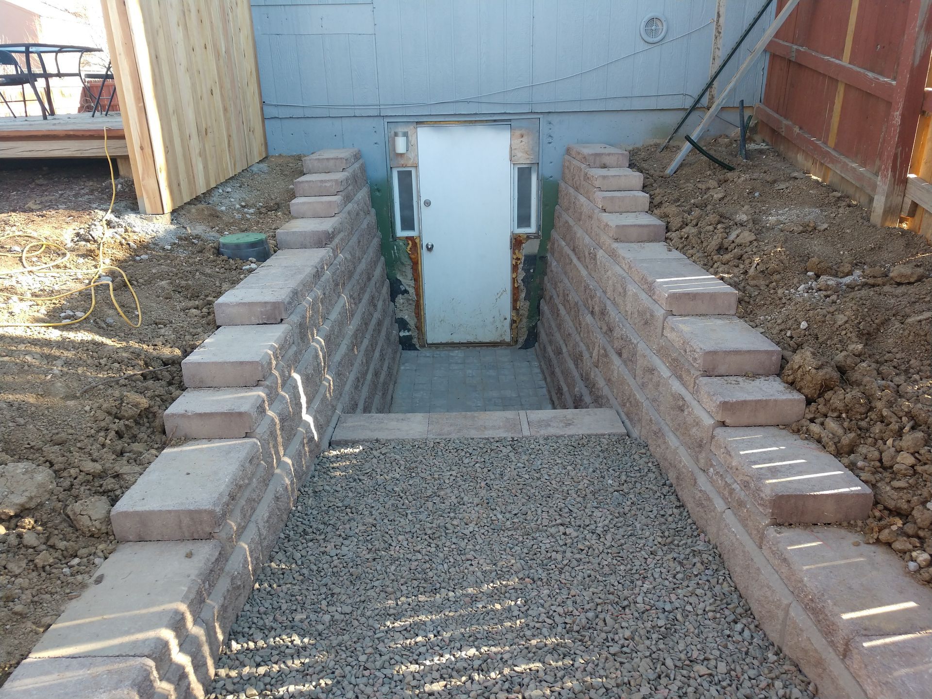 Stone-walled entryway leading to a basement door. Gray gravel covers the ground. Light brown retaining blocks line the walls.