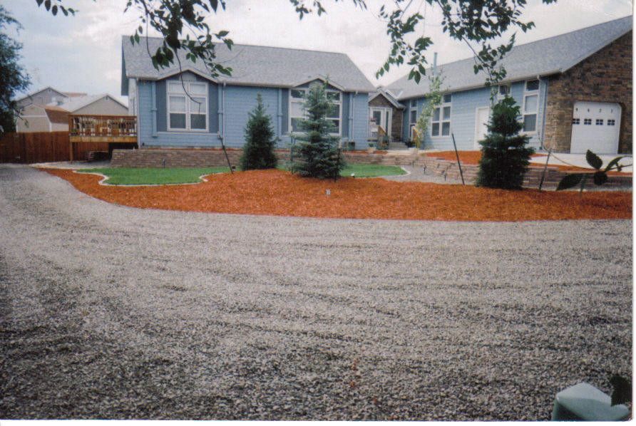 Blue house with a gravel driveway and a landscaped front yard with red mulch and small trees.