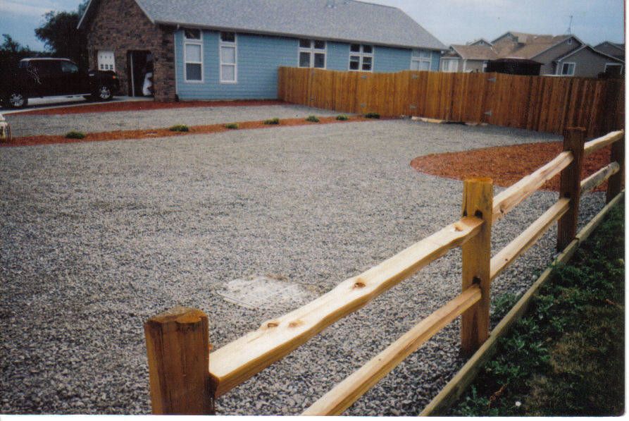 Gravel driveway with split-rail fence in the foreground, wooden fence around a blue house in the background.