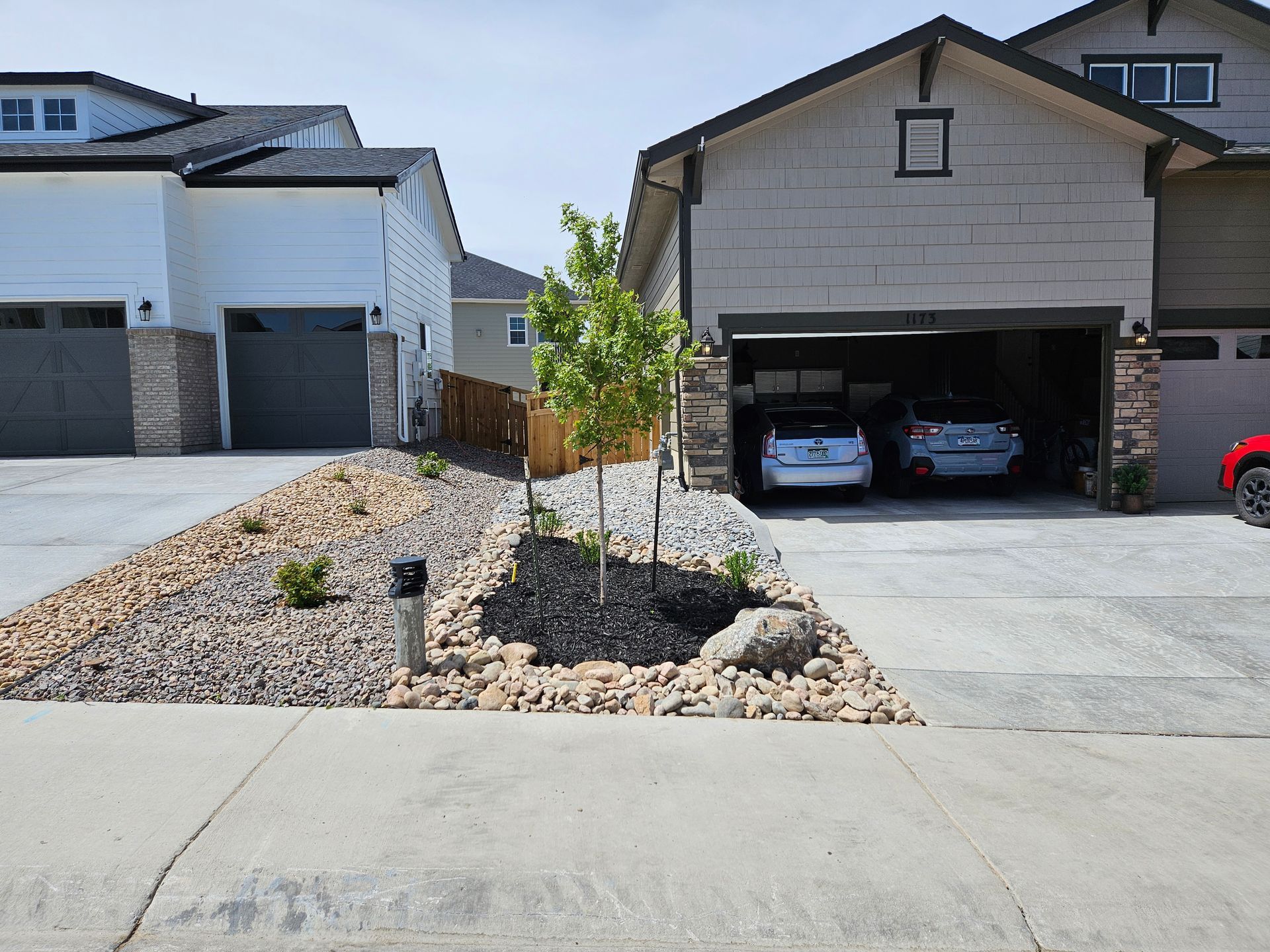 Suburban houses with attached garages. A landscaped yard features rocks and a small tree. Cars are parked in the open garage.