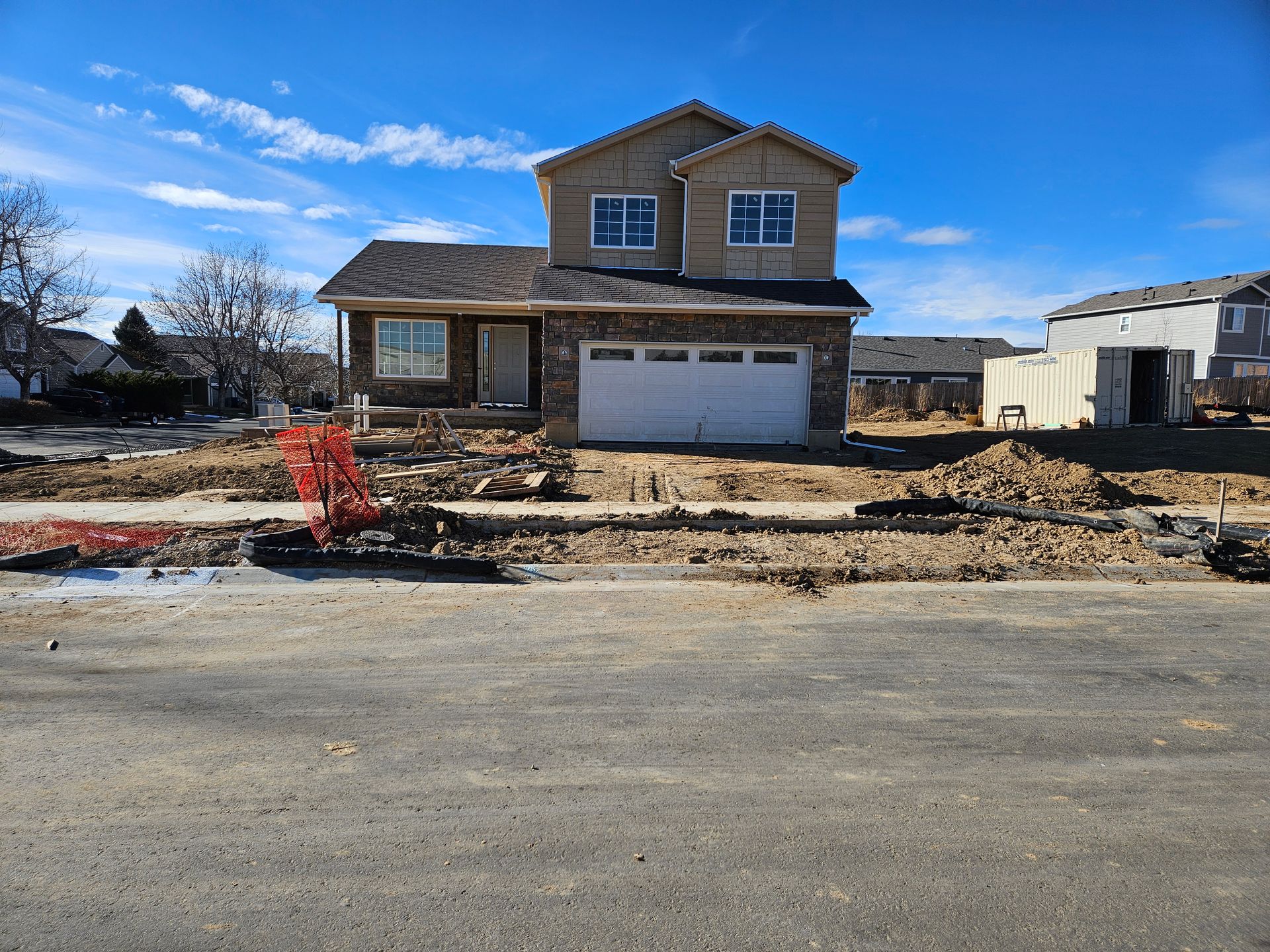New two-story house under construction with brown siding and stone accents on a sunny day. Front yard is dirt.