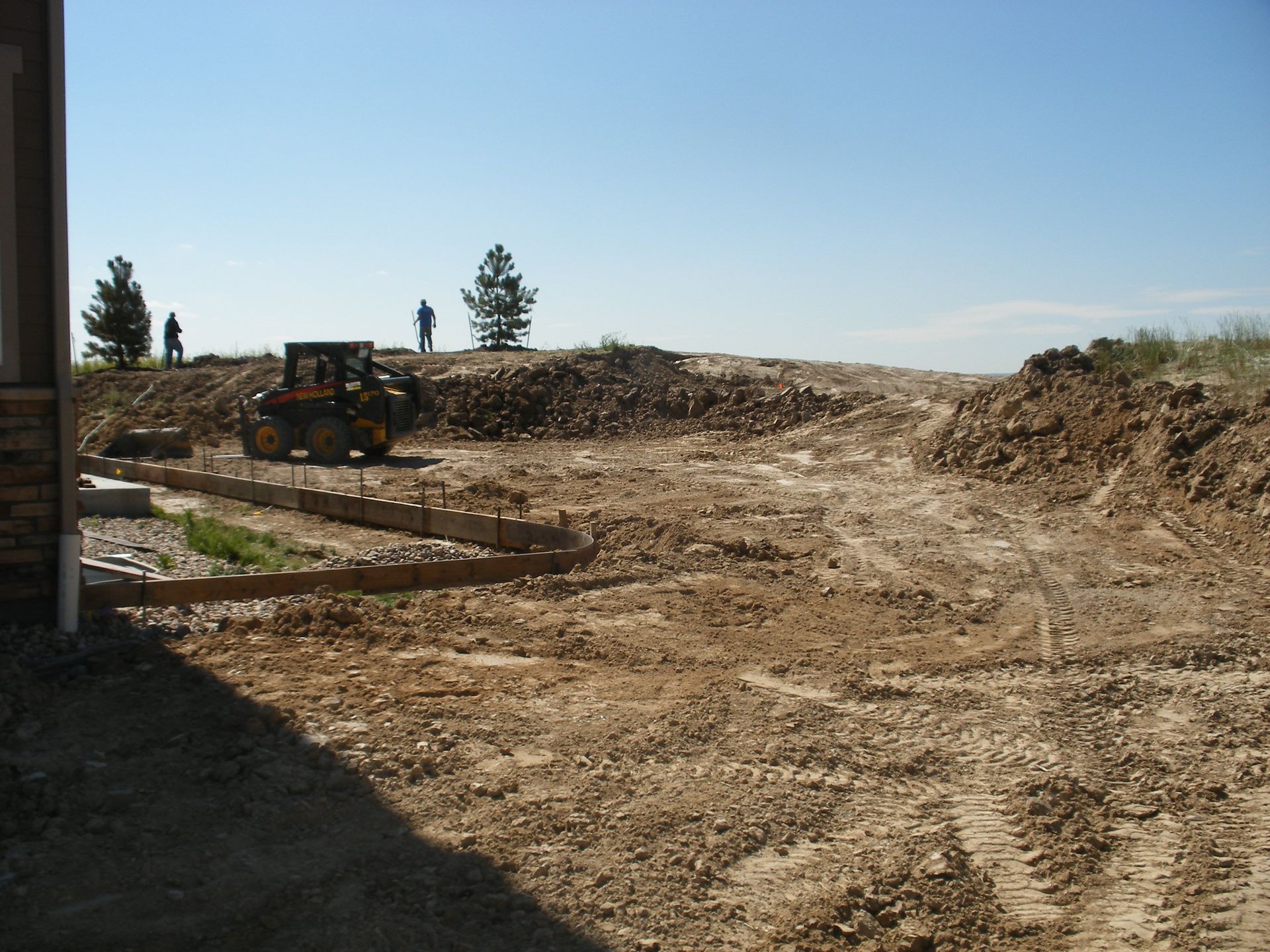 Construction site with dirt and gravel, a small bulldozer, and people standing on a mound under a blue sky.