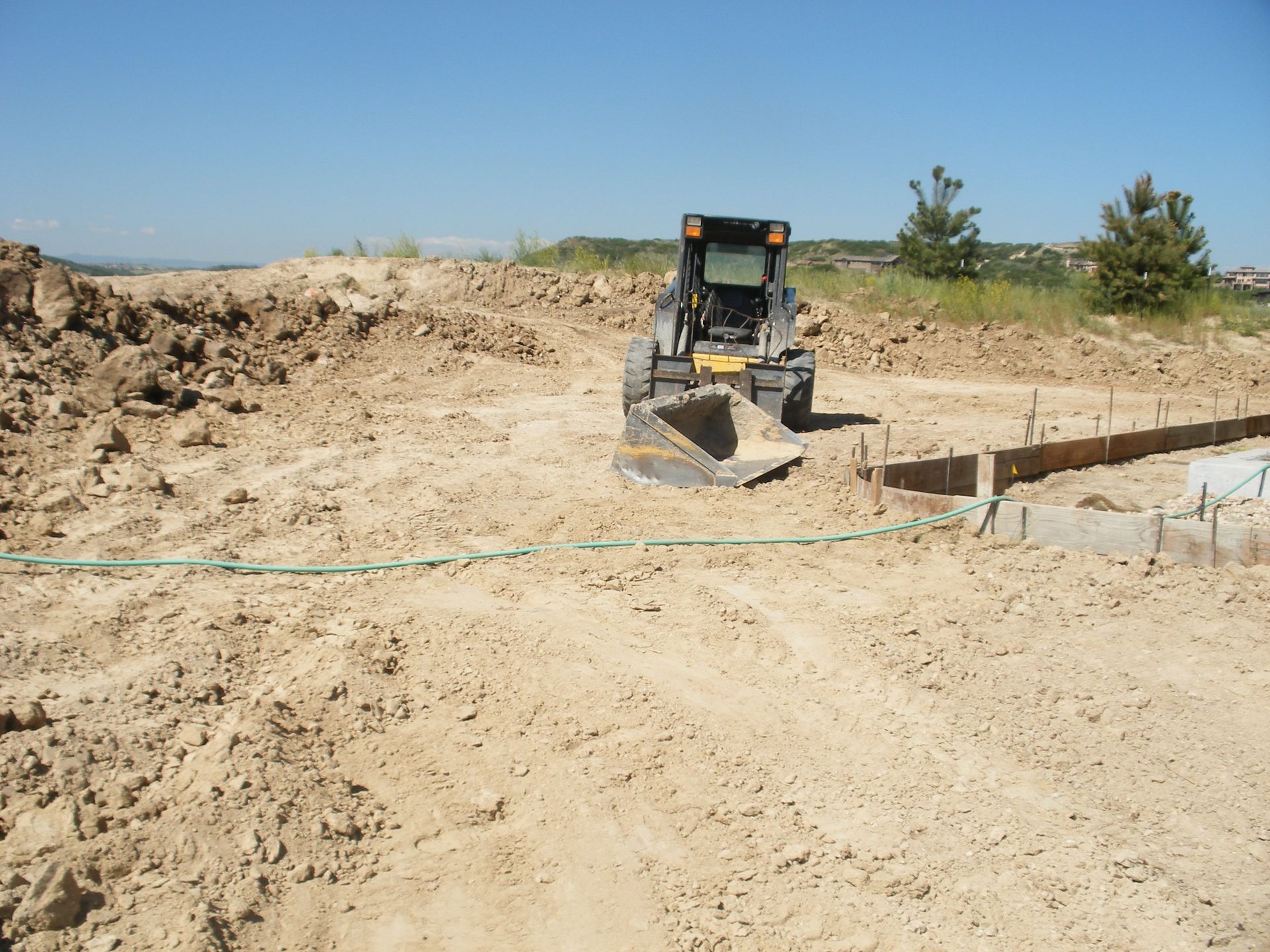 A small yellow and black skid steer loader moving sand on a construction site on a sunny day. Concrete forms are in the foreground.