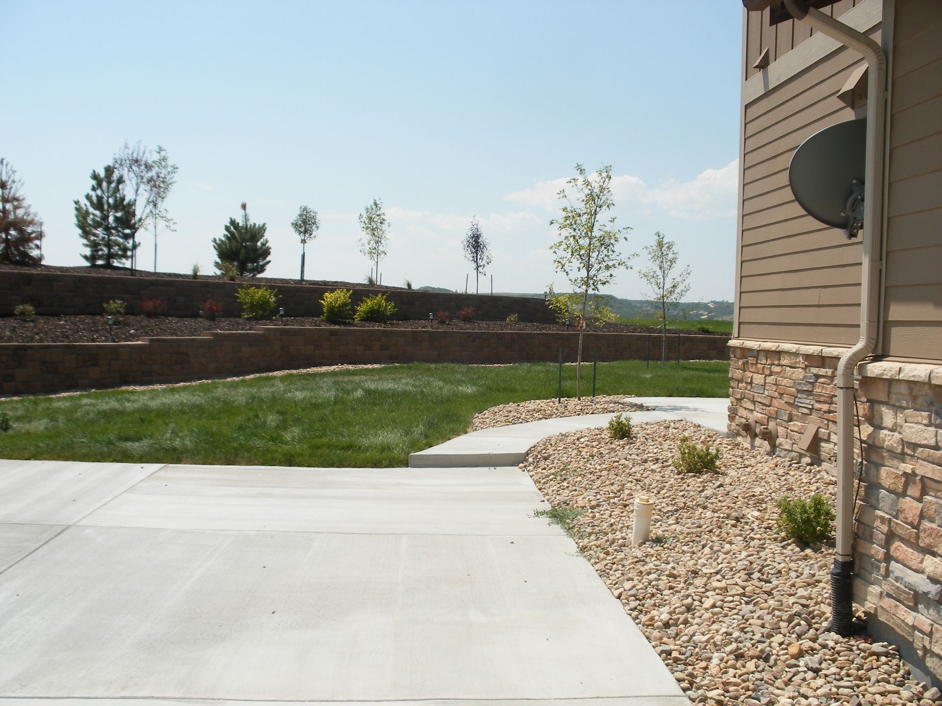A paved patio leads to a grassy lawn with rock landscaping. A stone wall and trees line the elevated background.