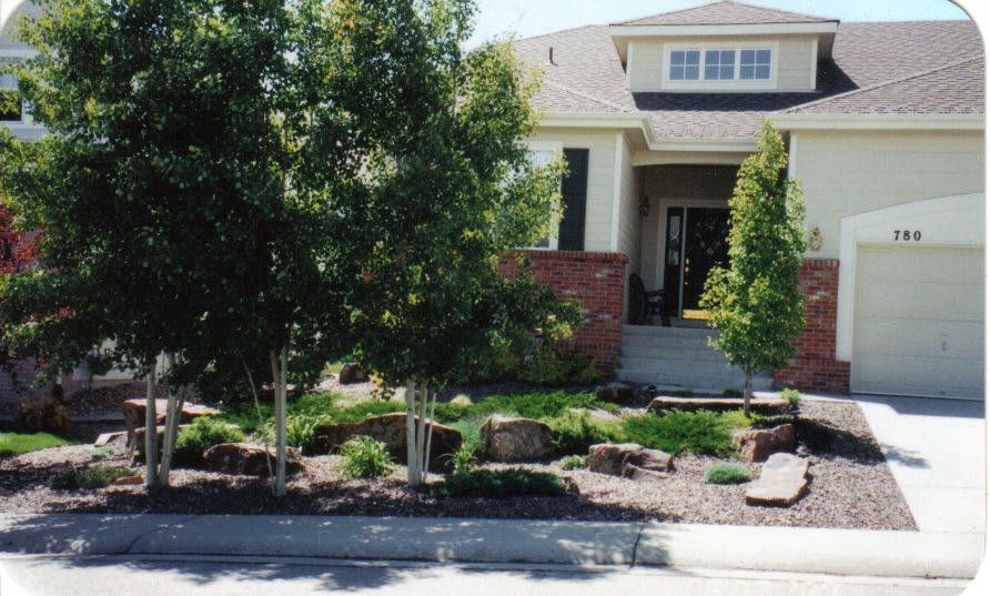 A house with a rock garden in the front. Trees and shrubs dot the landscape with a beige facade and a garage.