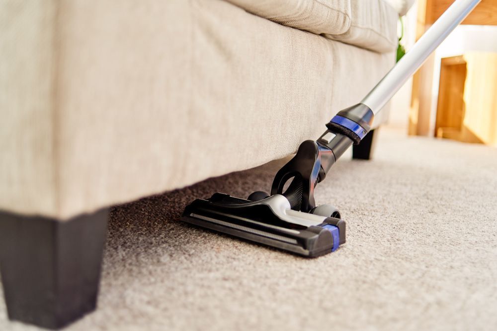 A Vacuum Cleaner Is Cleaning The Carpet Under A Couch In A Living Room — Echo Carpet Cleaning In Wauchope, NSW