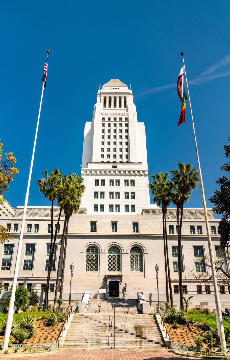 Los Angeles City Hall, a tall white building, with flags waving and palm trees under a blue sky.