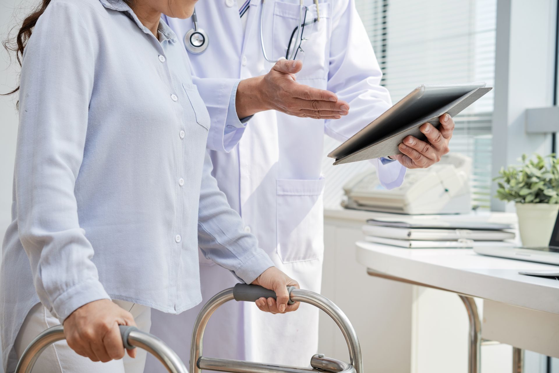 Woman using a walker consults with a doctor in a bright office; doctor holds a tablet, gesturing.