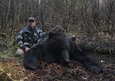 A man is sitting next to a large black bear in the woods.