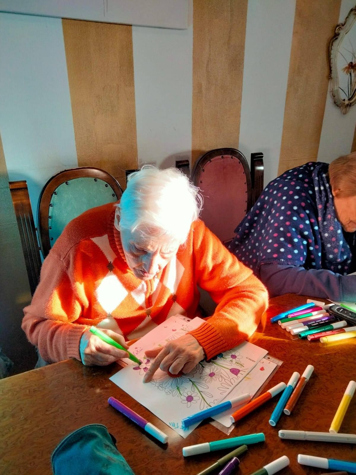 Una persona anziana con i capelli bianchi e un maglione arancione colora un foglio di carta con dei pennarelli seduti su un tavolo, illuminati dalla luce del sole. Un'altra persona è seduta lì vicino.