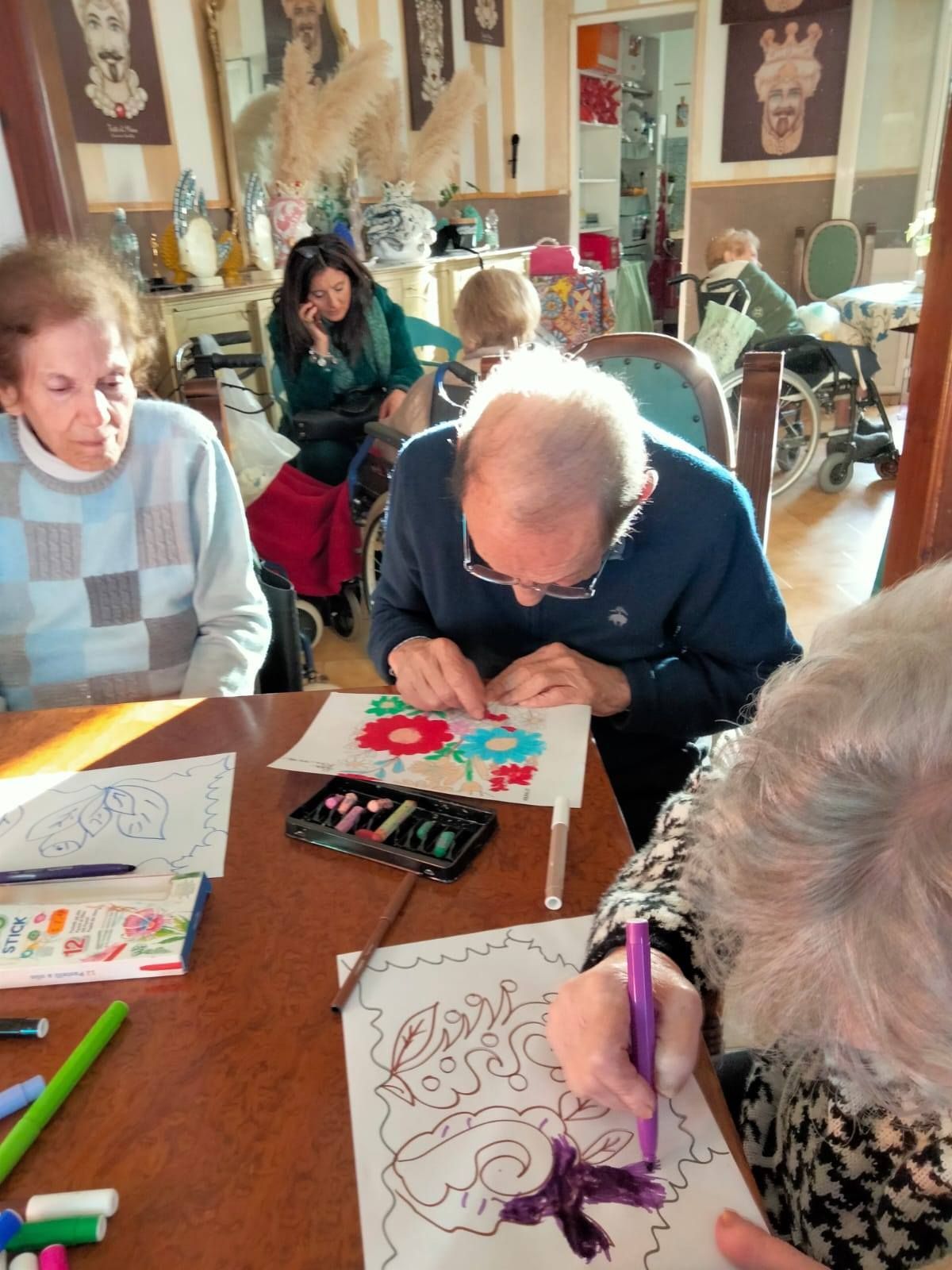 Un gruppo di anziani colora a un tavolo. Una donna con una camicia verde osserva dallo sfondo.
