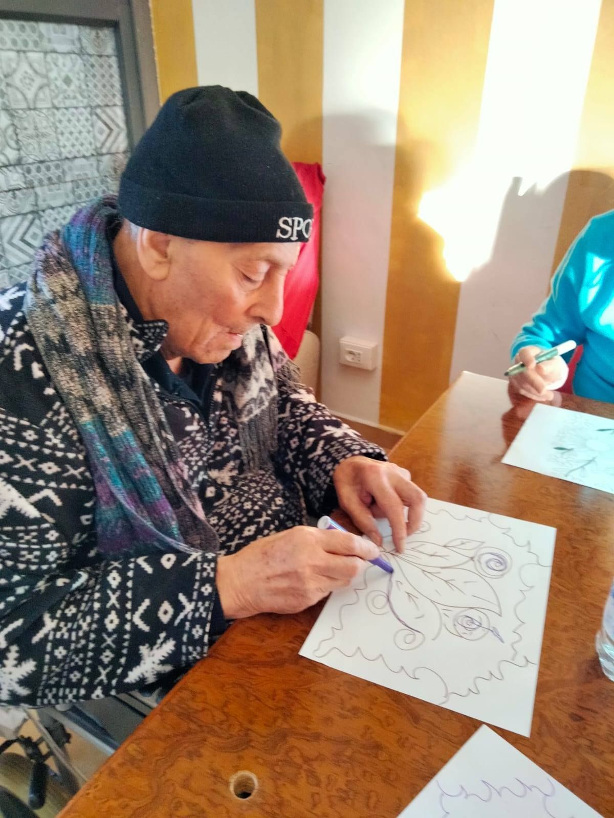 Una persona anziana, con indosso un cappello e una sciarpa, disegna su un foglio di carta con una penna, seduta a un tavolo, possibilmente in un contesto comunitario.
