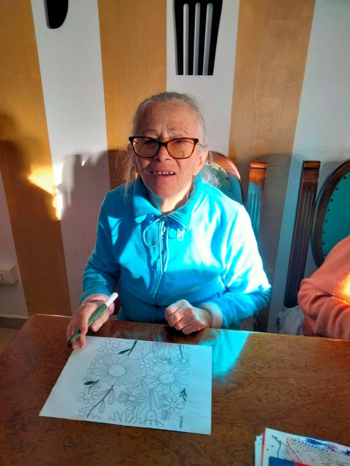 Una donna anziana con gli occhiali sorride, mentre colora un disegno floreale seduto a un tavolo di legno, in casa. Indossa una camicia blu.