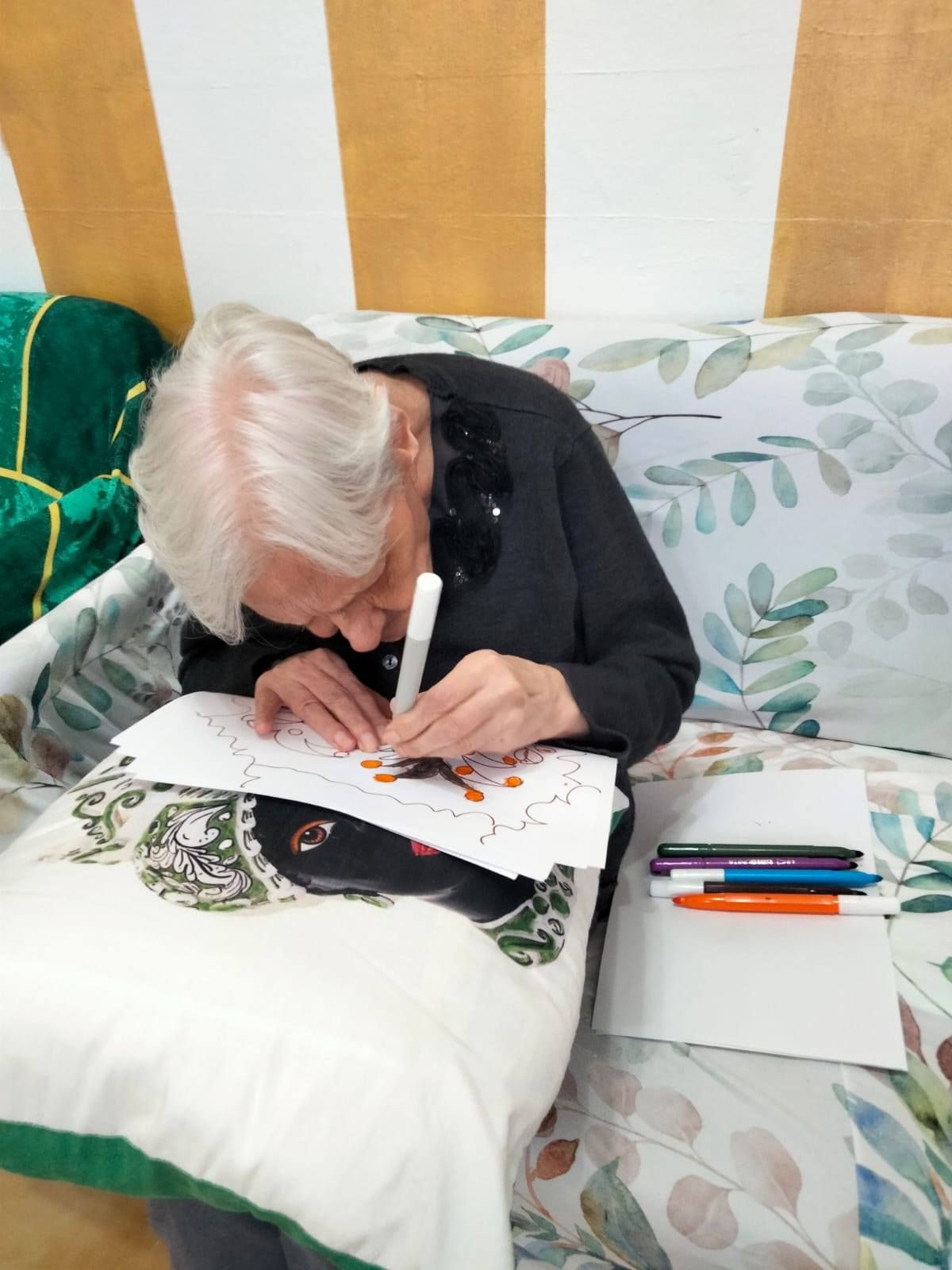 Una donna anziana con i capelli bianchi colora un disegno su un cuscino. È seduta su un divano e ci sono dei pennarelli lì vicino.