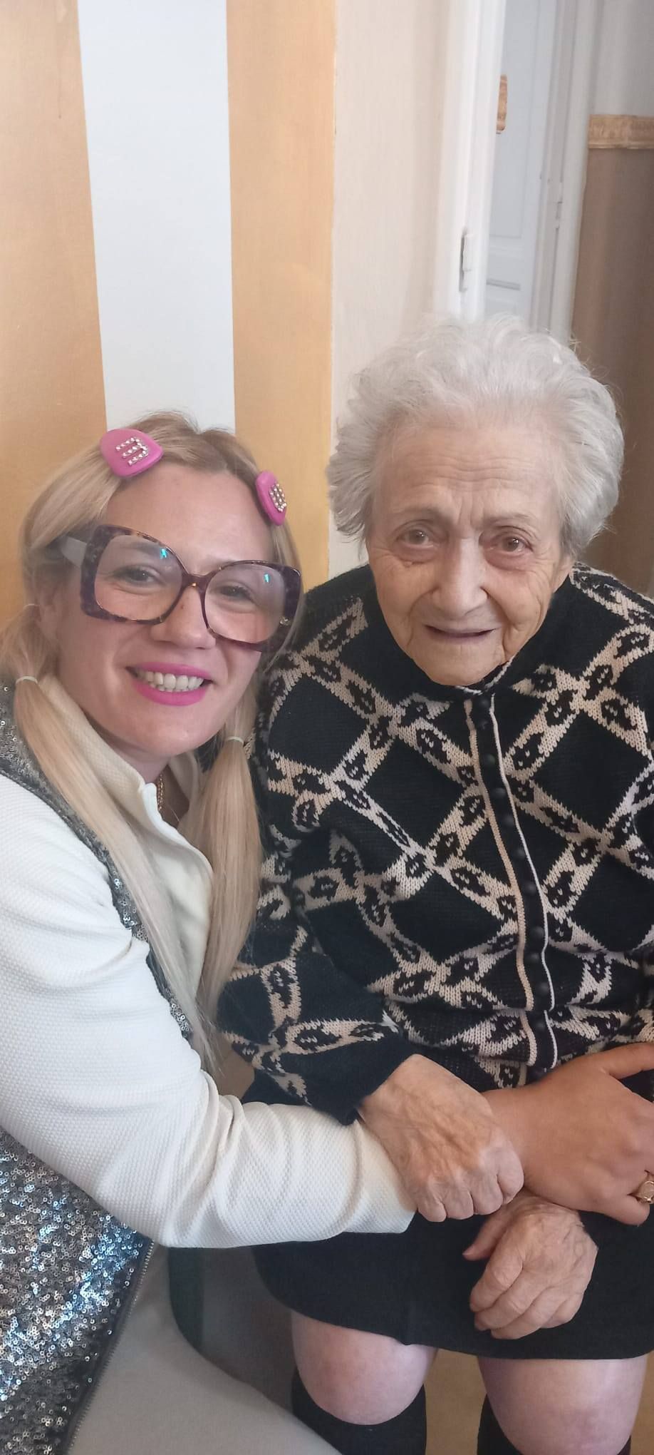 Due donne posano in un interno. Una, con grandi occhiali e un accessorio rosa per capelli, sorride. L'altra, una donna anziana, è seduta e guarda verso la telecamera.