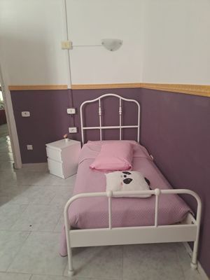Un piccolo letto di metallo bianco in una stanza con pareti viola e bianche. Il letto è dotato di lenzuola rosa e un cuscino a forma di panda.
