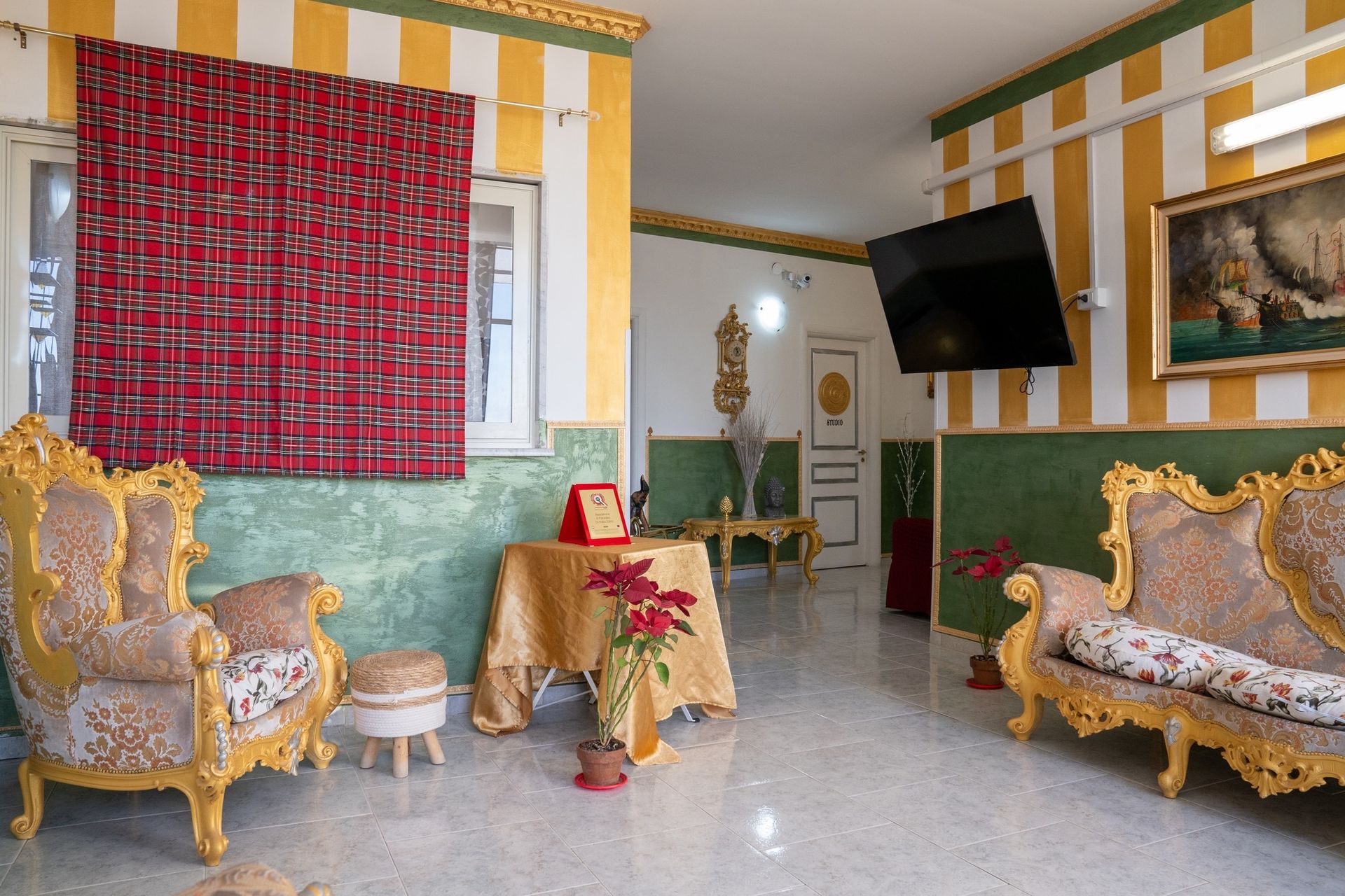 Soggiorno decorato con mobili con finiture dorate, paralume con motivi rossi e pareti a righe gialle e verdi. Un tavolo è apparecchiato con una composizione floreale rossa.