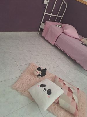 Una camera da letto rosa con un letto bianco, un soffice tappeto con cuscini e animali di peluche. Parete viola e pavimento piastrellato bianco.
