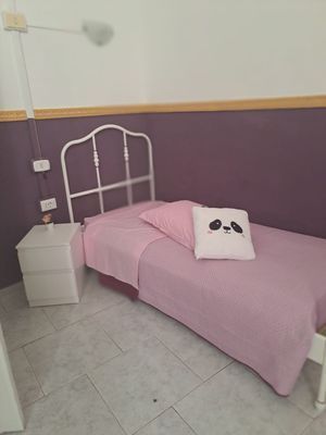 Una piccola camera da letto con un letto in metallo bianco, biancheria da letto rosa e un cuscino a forma di panda contro una parete viola.