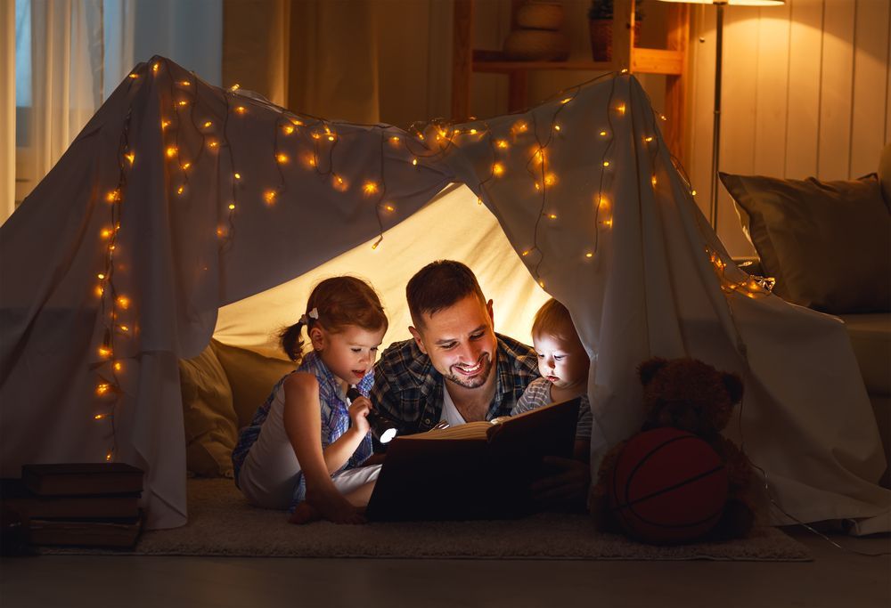 Father and daughters reading in pillow fort at night