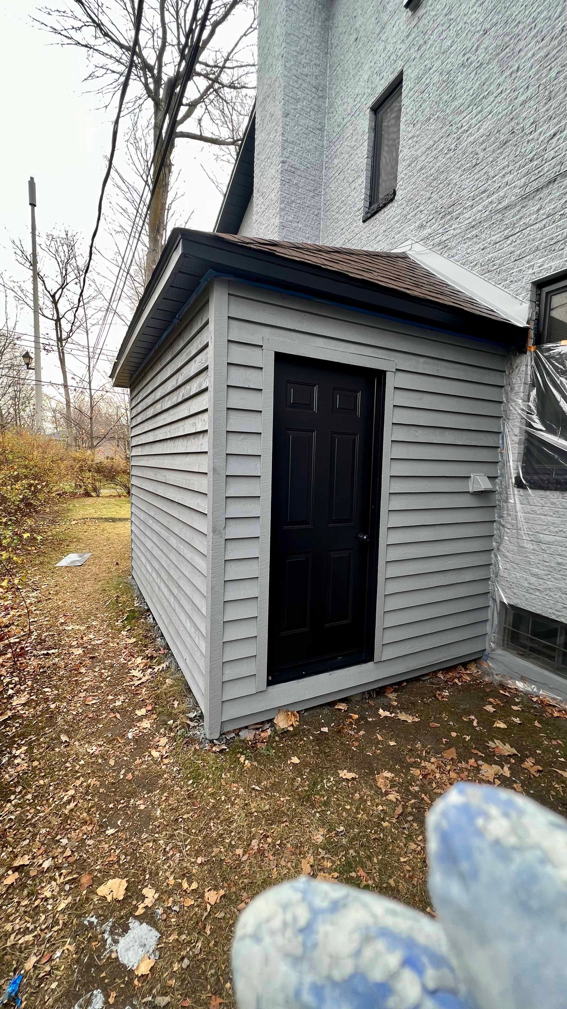 A small shed with a black door is in the backyard of a house.