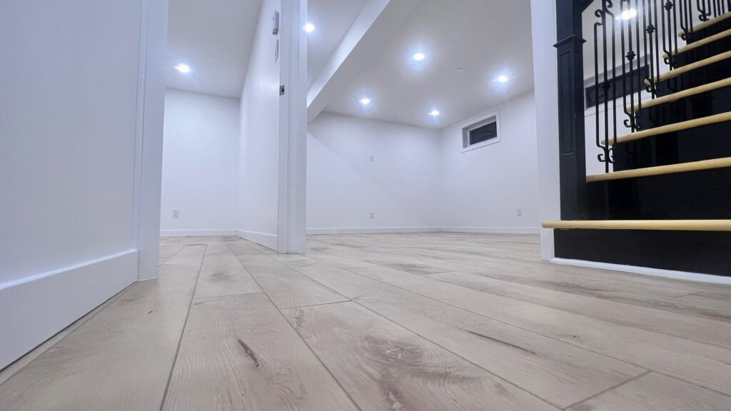 Brightly lit, empty basement room with light wood flooring and staircase.