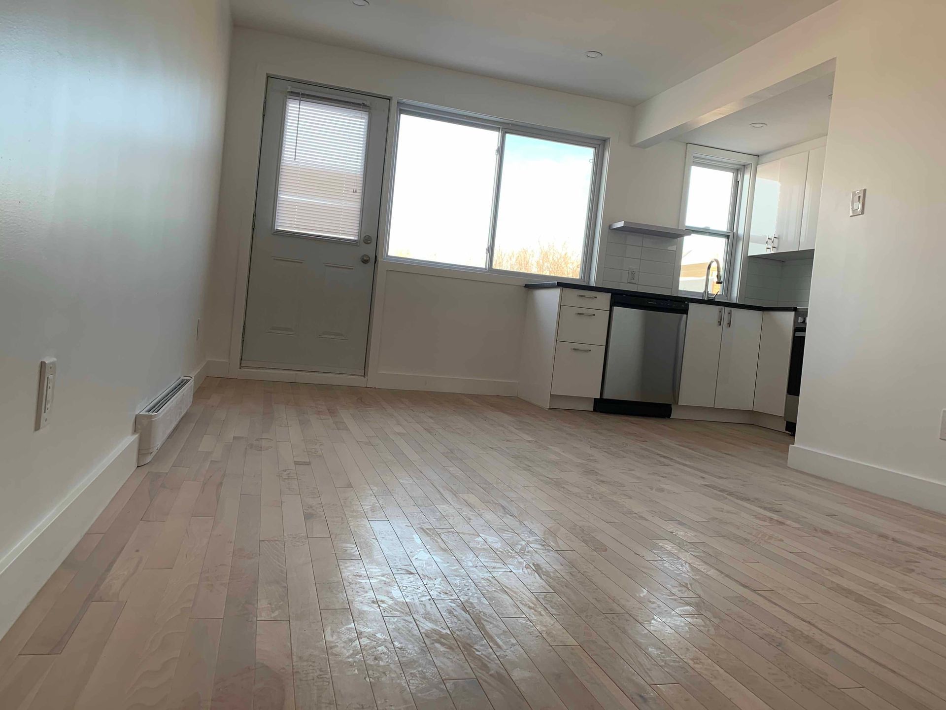 An empty room with hardwood floors and a kitchen in the background