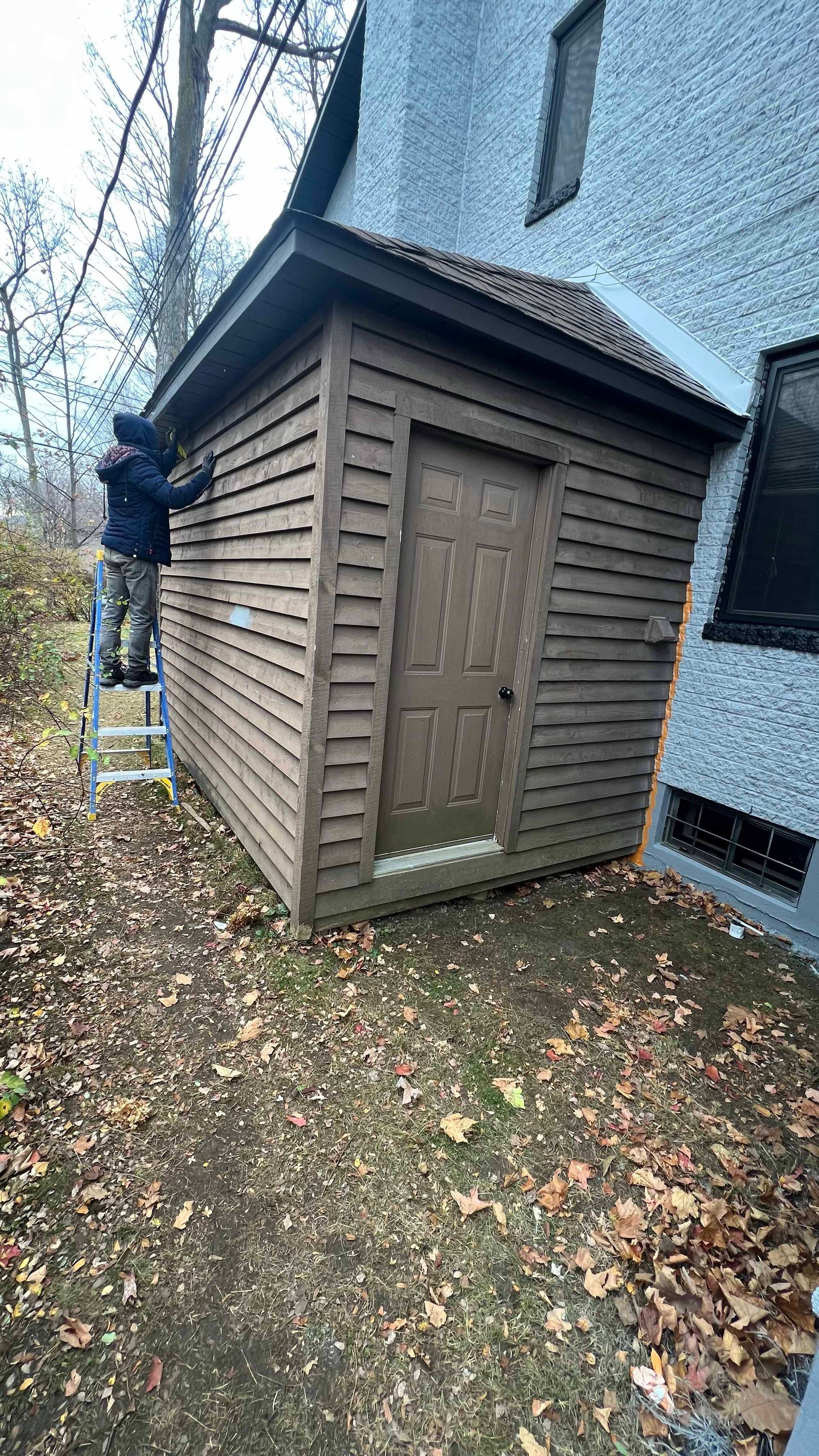 A man is standing on a ladder painting a shed in front of a house.