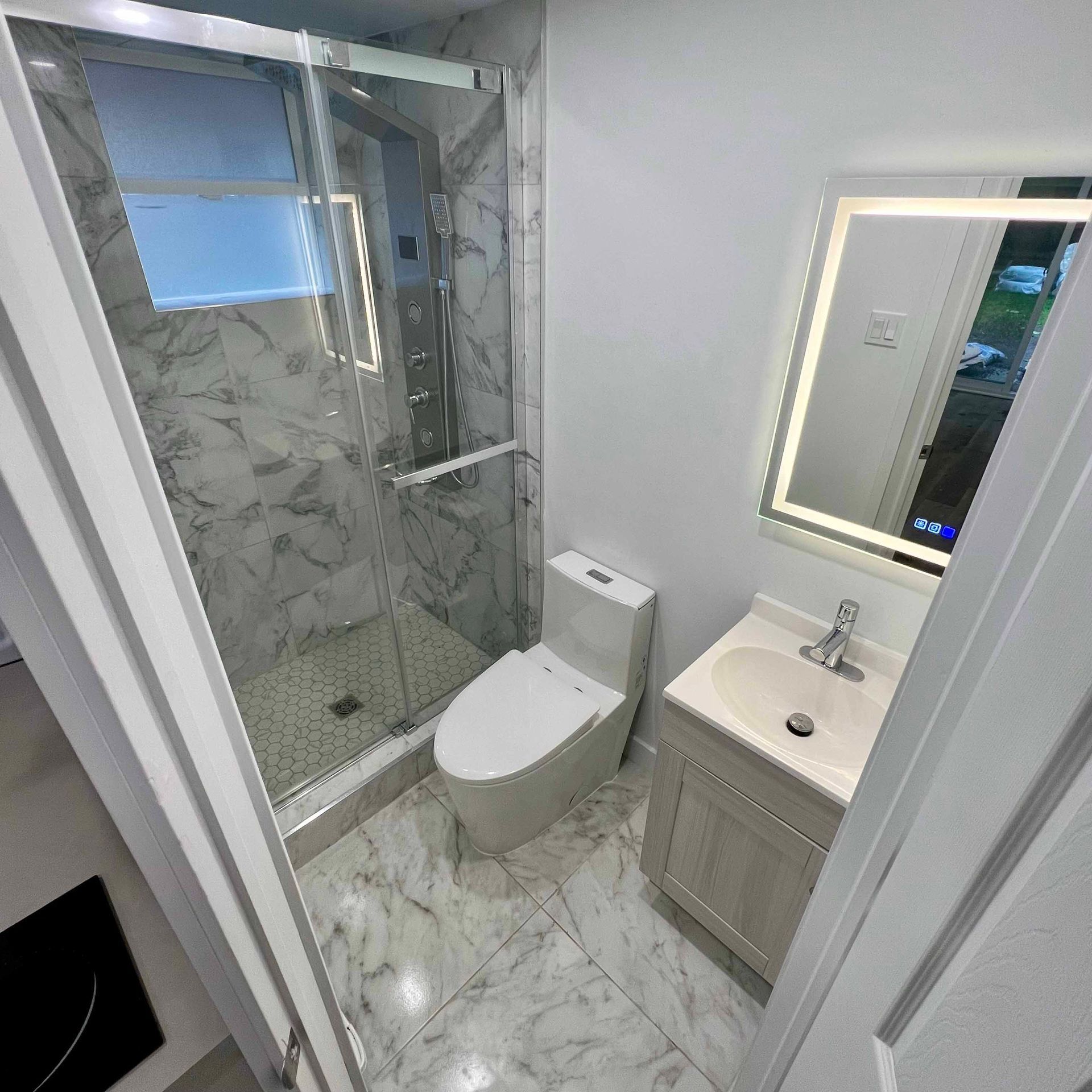Small, modern bathroom with marble tile, a glass shower, a toilet, and a vanity with a lighted mirror.