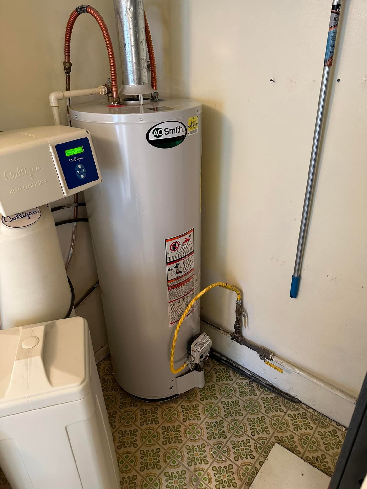 A white water heater is sitting in a room next to a mop.