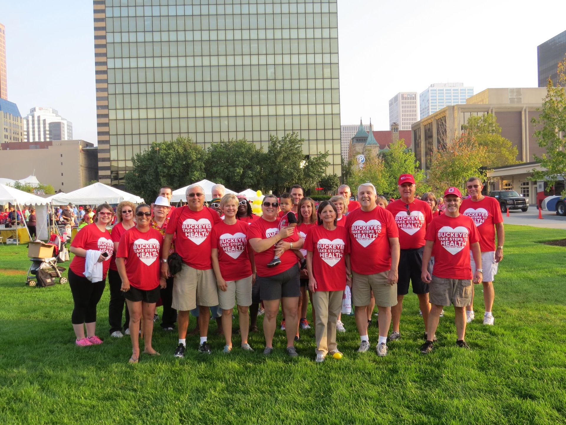 Group of people in red shirts, standing on grass, with buildings in the background.